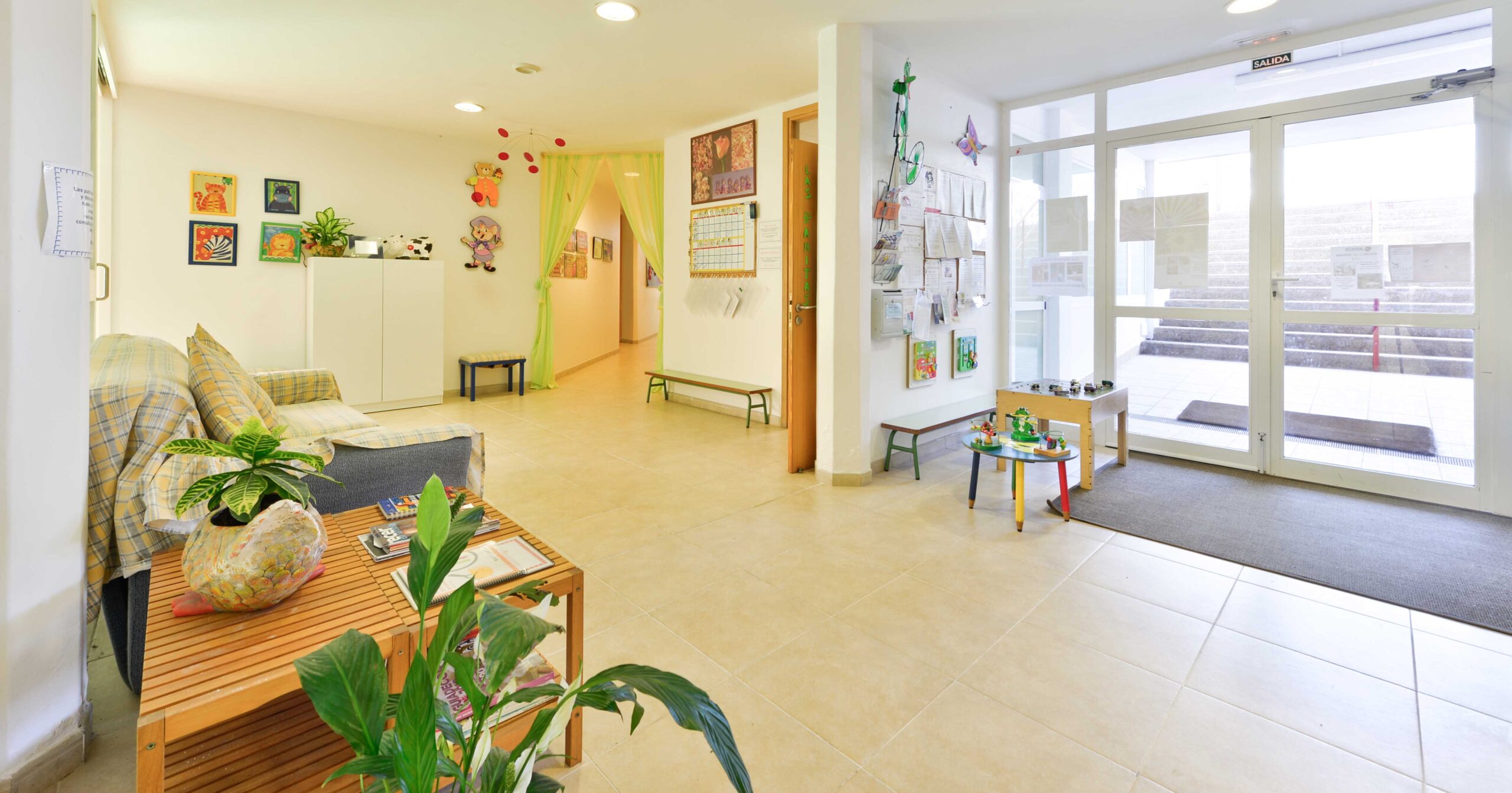Recepción luminosa y acogedora de una guardería o centro infantil, con decoración colorida, muebles sencillos y plantas. A la izquierda, un sofá con cojines y una mesa con libros y plantas. Al fondo, un pasillo decorado con dibujos y cortinas verdes. A la derecha, una puerta de cristal que da acceso al exterior y una zona de juegos con juguetes y mesas pequeñas.