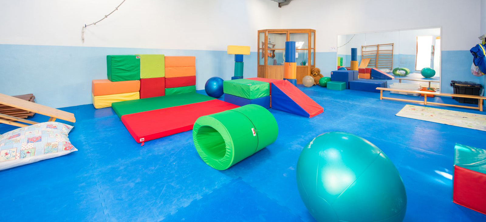 Espacio amplio y colorido de psicomotricidad en un centro infantil, con colchonetas, bloques de espuma de colores, pelotas grandes y túneles. El suelo es azul y las paredes tienen un diseño sencillo en tonos claros. El área está diseñada para fomentar el movimiento y el desarrollo motor de los niños.